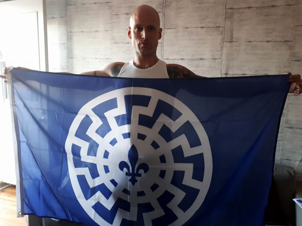  Philippe Gendron of the Soldiers of Odin showing off a made-to-order flag that combines the fleur de lys with the black sun, an occult Nazi symbol.